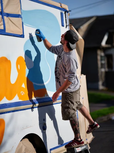 A tattooed muralist in a baseball cap and gray t-shirt reaches upward with a blue spray can, adding vibrant orange and blue elements to a white food truck's exterior. Sharp afternoon shadows dance across the vehicle's surface as the artist works methodically on the evolving mural. The urban scene unfolds on Portland's Northeast 9th Avenue, where residential architecture forms a soft backdrop to this creative transformation.
