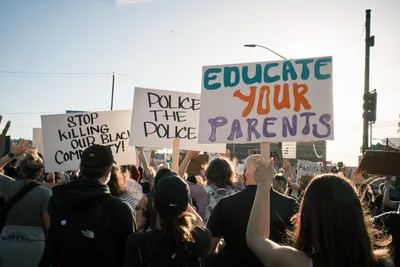 A dense crowd of protesters moves purposefully across Portland's Morrison Bridge, their handmade signs catching the golden hour light against a pale sky. The demonstrators hold messages including "STOP KILLING OUR BLACK COMMUNITY," "POLICE THE POLICE," and "EDUCATE YOUR PARENTS," their colorful lettering standing out against white poster board. The late afternoon sun backlights hair and shoulders, creating an atmosphere of determined solidarity as the group advances toward downtown Portland. Power lines and urban infrastructure frame the scene, emphasizing the grassroots nature of this civil rights demonstration.