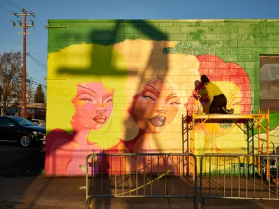 Multidisciplinary artist Villatose works from a scaffolding platform, adding finishing touches to 'Jardín Cubana,' a bold street mural featuring three women with flowing blonde hair rendered in brilliant yellows, oranges, and greens. The large-scale artwork dominates the concrete block wall on Northeast Alberta Street in Portland's historic Alberta Arts District, while golden hour light bathes the scene in warm, atmospheric tones. Safety barriers and urban infrastructure frame the creative process, capturing the intersection of art and community revitalization.