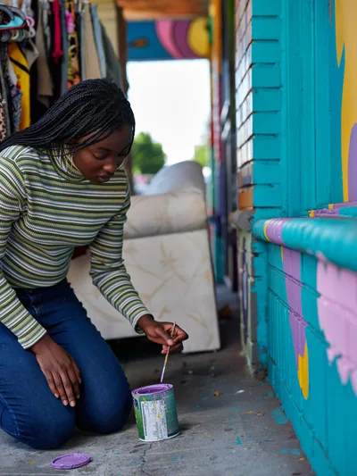 Naomi Likayi kneels on the sidewalk, carefully signing her vibrant turquoise and pink mural with purple paint in Portland's Alberta district. The intimate moment captures the artist's focused concentration as she puts the finishing touches on her colorful street art, surrounded by the creative energy of this Northeast Portland neighborhood. Natural daylight illuminates her striped sweater and braided hair as she works with steady precision, paint can beside her on the concrete.