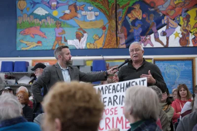 Senator Ron Wyden engages with constituents during a heated town hall meeting at Robert Gray Middle School in Portland's Hillsdale neighborhood. A bearded interviewer in a gray suit extends a microphone toward Wyden, who gestures emphatically while speaking to the packed audience. The scene unfolds against vibrant murals depicting community life, while protest signs calling for impeachment punctuate the charged political atmosphere. Rows of engaged citizens, many with graying hair, lean forward attentively in the school's multi-purpose room.