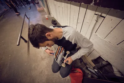 A young craftsman hunches over a skateboard deck in the industrial confines of Commonwealth Skateboarding on Southeast 20th Avenue in Portland's Buckman neighborhood. Warm overhead lighting casts dramatic shadows across weathered concrete floors as he meticulously adjusts the board's components, surrounded by the authentic clutter of skate tools and equipment. The intimate scene captures the meditative ritual of skateboard maintenance, where precision meets passion in this legendary Portland skate sanctuary.