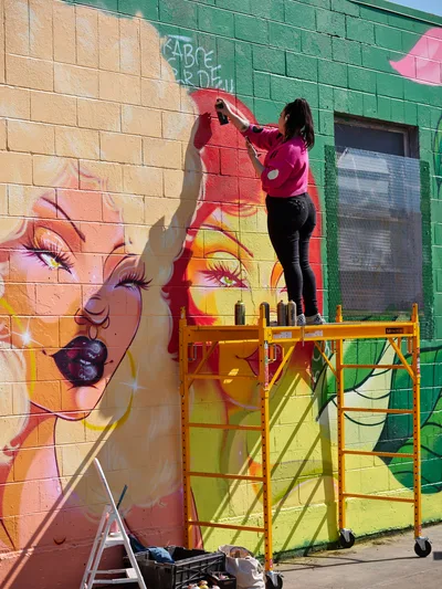 Multidisciplinary artist Vilatose works from a yellow scaffolding platform, spray painting vivid details on a large-scale portrait mural adorning a brick wall in Portland's Alberta Arts District. The emerging artwork features luminous skin tones, piercing green eyes, and bold red elements against the textured masonry canvas. Golden afternoon light casts dramatic shadows across the colorful work-in-progress, while paint supplies and equipment scatter below on the concrete sidewalk.
