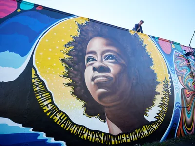 Travis Fields stands triumphantly atop the building beside his completed mural of poet Amanda Gorman in Portland's Alberta Arts District. The large-scale portrait depicts Gorman with a luminous golden halo framing her natural hair, her face rendered in warm earth tones with an upward gaze of hope and determination. The surrounding wall bursts with psychedelic swirls of magenta, cyan, and electric blue, creating a cosmic backdrop that celebrates both the subject and the vibrant street art culture of Northeast Portland.