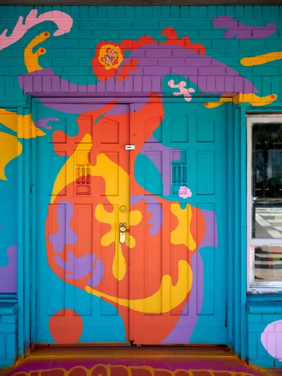 An abstract human figure dominates this explosive street mural by Naomi Likayi, painted across turquoise wooden doors and brick walls in Portland's Alberta Arts District. The character is adorned in flowing orange and purple garments, with wild hair rendered in kaleidoscopic hues against the cerulean backdrop. Bold geometric shapes and organic forms dance around the central figure, transforming an ordinary storefront entrance into a portal of artistic fantasy and neighborhood creative expression.