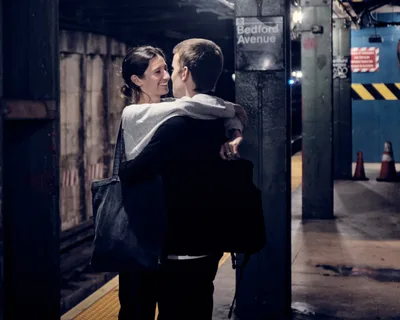 A tender moment unfolds on the platform of Bedford Avenue subway station in Williamsburg, Brooklyn, as a couple shares an intimate embrace beneath the station's iconic signage. The woman, wearing a light hoodie and carrying a dark bag, smiles warmly as she holds her partner dressed in dark clothing. The scene captures the raw authenticity of New York subway romance, with the station's industrial elements—weathered tiles, warning signs, and platform infrastructure—creating a gritty urban backdrop bathed in the station's harsh fluorescent lighting.
