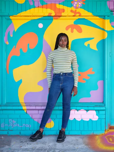 Naomi Likayi stands confidently before her completed mural on Northeast 15th Avenue in Portland's Alberta district. The young Black artist wears a striped turtleneck and high-waisted jeans against the explosive backdrop of turquoise, yellow, orange, and purple abstract shapes that dance across the storefront's surface. Natural daylight illuminates both subject and artwork, creating a harmonious composition that celebrates urban art and creative expression in this gentrifying Portland neighborhood.