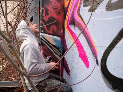 Travis Fields, known as Campographic, crouches beside a concrete wall at Clay Creative Building in southeast Portland's Hosford-Abernethy neighborhood, spray can in hand as he works on a bold mural. The artist, wearing a light gray hoodie, gazes upward at his evolving creation of flowing magenta, orange, and crimson forms against weathered concrete. Dormant winter grasses frame the intimate creative moment, capturing the meditative focus of urban artistry in progress.