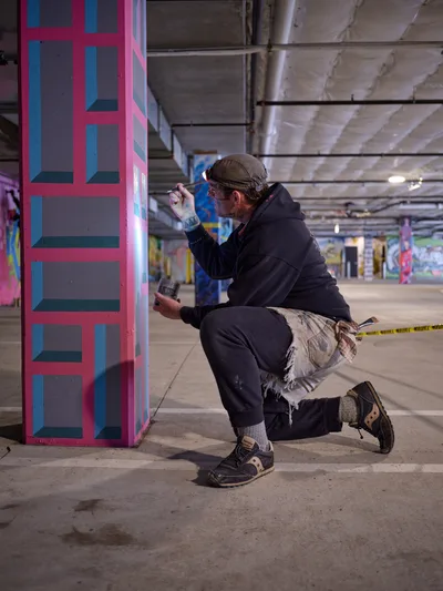 Bike activist and artist Rabbitt kneels in focused concentration as he applies precise brushstrokes to a vibrant geometric mural on a concrete column in the Clay Creative building's parking garage. The underground space, illuminated by harsh fluorescent lighting, transforms into an impromptu studio where pink and blue angular patterns emerge against the industrial backdrop. His paint-stained work clothes and methodical approach capture the dedication required to bring color to Portland's urban spaces.