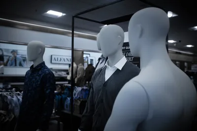 Three mannequins stand in stark contrast within Nordstrom's Clackamas location, their featureless forms creating an unsettling tableau under harsh fluorescent lighting. Two figures are impeccably dressed in professional attire—one in a navy patterned shirt, another in a charcoal vest with crisp white collar—while a third stands naked, its pale form catching the light like an uninvited guest at a formal gathering. The scene captures the surreal intersection of commerce and humanity, where plastic bodies serve as silent ambassadors for fashion consumption.