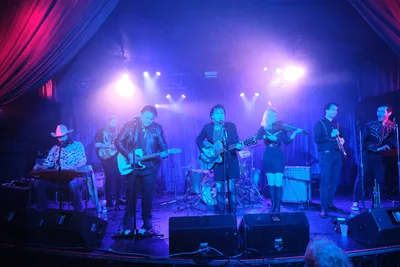 Six-piece band Roselit Bone performs under dramatic blue and purple stage lighting at the intimate Dante's venue in Portland, Oregon during RX Fest 2019. The atmospheric concert photography captures the musicians mid-performance, with the lead vocalist at center microphone flanked by guitarists, a violinist, and a keyboardist in cowboy hat. Theatrical lighting cuts through hazy air, creating a moody underground club aesthetic framed by burgundy curtains.
