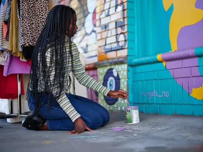 Naomi Likayi kneels on concrete pavement as she carefully signs her vibrant mural on a turquoise and purple brick wall in Portland's Alberta neighborhood. Her long braided hair falls forward as she concentrates on her brushwork, wearing a striped long-sleeve shirt and blue jeans. The colorful street art backdrop includes clothing displays and urban elements that speak to the area's creative energy. Warm natural light illuminates this moment of artistic completion, capturing the intimate act of an artist claiming her work.