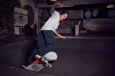A tattooed skateboarder in white shirt and dark pants carves through a concrete bowl at Commonwealth Skateboarding in Portland's Buckman neighborhood. The underground venue's raw industrial atmosphere is punctuated by graffiti-covered walls, exposed beams, and skull artwork, creating a gritty backdrop for this editorial skateboarding session. Warm lighting illuminates the rider's focused expression as he navigates the curved concrete surface.