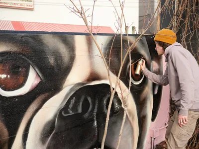 Muralist Wetiko applies golden ochre spray paint to an expressive portrait mural at Clay Creative in Portland's Hosford-Abernethy neighborhood. Wearing a mustard beanie and gray hoodie, the artist works with focused precision against the wall's dramatic black and flesh-toned imagery. Bare winter branches frame the scene, creating natural shadows that dance across the evolving artwork in the afternoon light.
