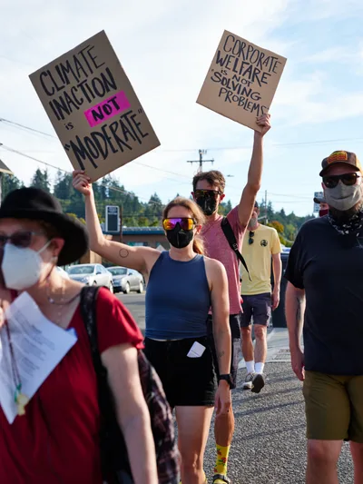 Masked protesters stride purposefully down McLoughlin Promenade in Oregon City, their handmade cardboard signs declaring "Climate Inaction is NOT Moderate" and "Corporate Welfare Not Solving Problems" against a backdrop of power lines and suburban storefronts. The afternoon sun casts sharp shadows across the asphalt as the determined group advances toward Representative Kurt Schrader's office. Their reflective sunglasses and face coverings create an anonymous solidarity while colorful athletic wear adds vibrancy to this grassroots demonstration for environmental action.