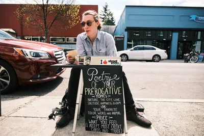 Till Gwinn, a local street poet, sits with legs spread apart behind his vintage typewriter on Portland's Alberta Street, putting finishing touches on a custom poem for a customer. His chalkboard sign advertises 'Poetry of Your Prerogative Topics' with handwritten details about pricing and payment options. The afternoon light bathes the scene as cars pass by on this bustling Northeast Portland thoroughfare, capturing the intersection of urban life and creative expression in one of the city's most vibrant neighborhoods.