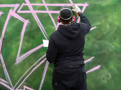 A muralist in black hoodie and baseball cap works intently on a dynamic pink and green graffiti piece at Clay Creative in Southeast Portland's Hosford-Abernethy neighborhood. The artist reaches upward with spray can in hand, adding flowing pink outlines to an emerald green background that dominates the wall surface. Red headphones frame their focused concentration as they craft bold geometric patterns and sweeping letterforms across the urban canvas.
