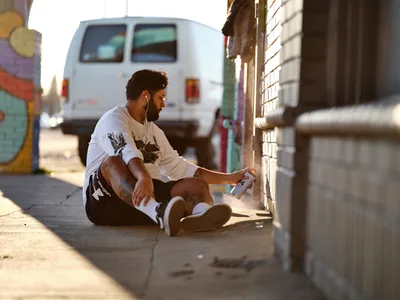 Muralist Heysus sits cross-legged on the concrete sidewalk in Portland's Alberta Arts District, carefully working on a wall mural as golden hour light illuminates his concentrated form. The artist, wearing a white graphic t-shirt and dark shorts, holds spray paint or markers while positioned against weathered brick, with a white van and colorful street art visible in the soft-focused background. The warm, directional lighting creates dramatic shadows and highlights the intimate moment of artistic creation in this vibrant Northeast Portland neighborhood.