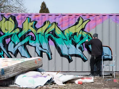 A graffiti artist in black clothing applies finishing touches to a dynamic wildstyle piece featuring flowing turquoise and lime green letters against a purple-pink background. The scene unfolds on a corrugated metal fence in Northeast Portland, Oregon, with scattered paint-stained tarps and a step ladder indicating an active work session. Bare winter trees and overcast skies frame this urban art creation, capturing the raw energy of street culture and creative expression.