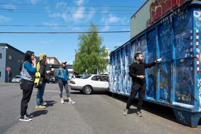 Tomás Valladares of the Portland Street Art Alliance gestures toward vibrant blue graffiti adorning a dumpster while leading an engaged group of young adults through Portland's Central Eastside. The afternoon scene captures the intersection of grassroots cultural education and urban expression, as participants listen attentively against a backdrop of industrial buildings and power lines. Sunlight illuminates the weathered asphalt and eclectic mix of street art that transforms utilitarian infrastructure into canvases of creative rebellion.