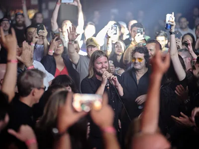 Band of Skulls frontman Russell Marsden grips the microphone with infectious energy as he performs at Webster Hall's intimate stage, surrounded by a sea of glowing smartphone screens held aloft by devoted fans. The atmospheric lighting cuts through the haze, illuminating his long blonde hair and genuine smile while capturing the electric connection between performer and audience. Behind him, bandmate Emma Richardson shares in the moment, while the packed crowd creates a constellation of digital documentation in the historic Manhattan venue.