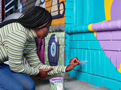 Naomi Likayi kneels beside her vibrant mural on Alberta Street, carefully applying her signature with a fine brush to the electric blue section of the painted brick wall. Her intricately braided hair cascades as she leans into her work, wearing a striped long-sleeve top that echoes the rhythmic patterns of her street art. Behind her, the colorful geometric mural stretches across the wall in bold turquoise, purple, and yellow blocks, transforming the urban corner into a canvas of community expression in Portland's historic Alberta Arts District.
