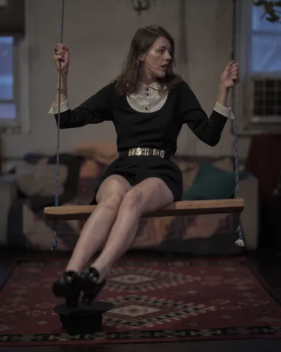 A young woman sits pensively on a wooden swing suspended in the warm interior of a Greenpoint apartment. Her dark dress contrasts with the cream-colored blouse beneath, while golden belt hardware catches the ambient light. The rope swing, weathered wooden seat, and patterned rug below create an intimate domestic tableau, with soft window light filtering through the Brooklyn space.