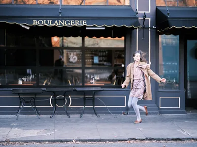A young woman in motion hurries past a charming French bakery on Portland's Alberta Street, her camel coat and plaid dress flowing with the breeze. The classic navy blue storefront with traditional awning and wrought iron chairs creates an intimate European café atmosphere. Warm afternoon light illuminates the scene as she strides purposefully along the sidewalk, embodying the energy of urban life in this trendy neighborhood.