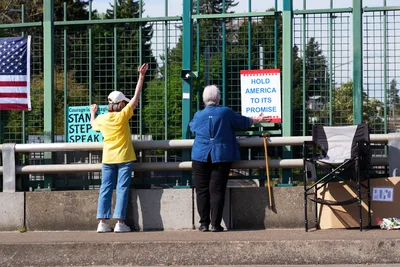 Two older activists prepare protest signage along the North Portland overpass above Interstate 5, their weathered hands gripping messages of resistance against the chain-link barrier. Morning light filters through the industrial mesh fencing as one woman in a bright yellow shirt raises her arm while her companion in denim secures a "Hold America to Its Promise" banner. Their determined silhouettes frame a moment of grassroots activism in the shadow of the Humboldt neighborhood's concrete infrastructure.