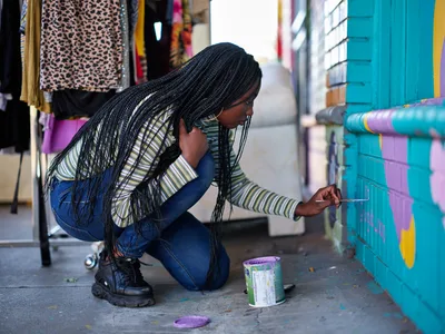 Naomi Likayi crouches in concentrated focus as she delicately signs her completed mural on a turquoise wall in Portland's vibrant Alberta Arts District. Her long black braids cascade forward as she works, paintbrush in hand, with a can of purple paint at her side. The urban street scene behind her reveals clothing displays and colorful storefronts, capturing the creative energy that defines this Northeast Portland neighborhood.