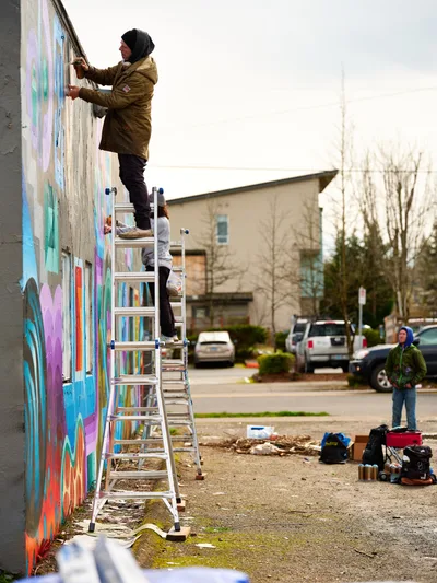 A hooded artist in an olive-green jacket stands on an aluminum ladder, spray painting a vibrant mural across a concrete wall in Northeast Portland, Oregon. The kaleidoscope of blues, purples, and warm tones transforms the urban facade while another observer watches from the sidewalk. Scattered art supplies and cans dot the pavement below, capturing the authentic process of community street art creation on an overcast day.
