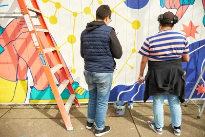 Two artists work intently on a vibrant public mural titled "Let's Talk" at Open Signal in Portland's historic Eliot neighborhood. The scene captures the collaborative creative process as Anke Gladnick and Maria Rodriguez paint geometric patterns and bold colors across the white wall. An orange ladder stands ready while paint buckets rest on the concrete sidewalk, with the emerging artwork featuring a dynamic interplay of yellow nodes, blue elements, and coral-toned forms that speak to community connection and dialogue.