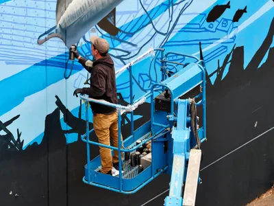 Street artist Jeremy Nichols commands a blue scissor lift against the stark concrete wall of Southeast Portland's Central Eastside district, his spray can releasing a cascade of azure tones across an emerging large-scale mural. Golden afternoon light bathes the industrial corridor of 2nd Avenue as the artist, clad in mustard work pants and brown hoodie, transforms the urban landscape into a flowing dreamscape of cerulean waves and abstract forms. The mechanical precision of the lift contrasts beautifully with the organic fluidity of his artistic vision, capturing the intersection of industrial infrastructure and creative expression that defines Portland's vibrant street art scene.