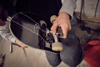 In the workshop atmosphere of Commonwealth Skateboarding on Southeast 20th Avenue in Portland's Buckman neighborhood, skilled hands make precise adjustments to a black skateboard's truck assembly. The intimate overhead perspective captures the mechanical intimacy of skate maintenance, with weathered concrete floors and workshop tools creating an authentic backdrop. Warm, diffused lighting bathes the scene in golden tones, emphasizing the tactile relationship between craftsperson and equipment in this celebrated Portland skate shop.