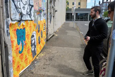 Tomás Valladares of the Portland Street Art Alliance guides participants through the vibrant wheat paste murals adorning concrete walls along Southeast Yamhill Street. Two men examine the layered urban artwork featuring bold yellow damask patterns, blue elephant imagery, and monochromatic character studies that transform the industrial corridor. The overcast Pacific Northwest light creates a muted atmosphere as the group moves through this celebrated gallery of sanctioned street art in Portland's evolving Central Eastside district.