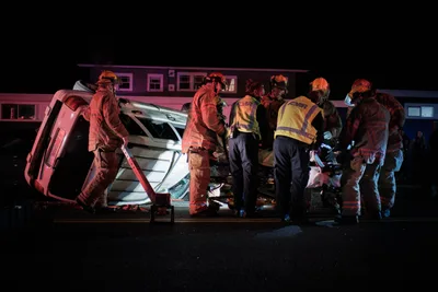Portland firefighters and paramedics work urgently around an overturned silver SUV on Northeast Killingsworth Street in the Humboldt neighborhood. The scene is bathed in dramatic red and blue emergency lighting against the stark black night, creating an intense atmosphere of coordinated rescue efforts. Multiple first responders in high-visibility gear and protective helmets cluster around the damaged vehicle, their movements deliberate and focused as they tend to injured passengers. The residential backdrop of modest homes provides a sobering contrast to the chaos of twisted metal and emergency response.