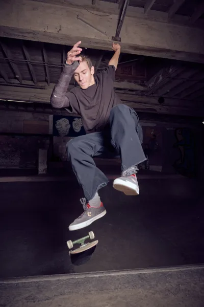 A young skateboarder hovers in mid-air beneath the weathered wooden beams of Commonwealth Skateboarding in Portland's Buckman neighborhood, his body frozen in graceful suspension above his board. The dramatic lighting cuts through the shadows of this basement skate haven, illuminating his focused expression and relaxed posture against the raw industrial architecture. The composition captures the poetry of skateboarding culture—where gravity momentarily surrenders to skill and style.