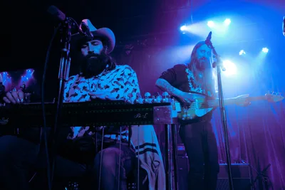 Barry Walker and Tobias Berblinger of Roselit Bone command the intimate stage at Dante's in Portland, bathed in ethereal blue and purple stage lighting. Walker, sporting a wide-brimmed hat and patterned shirt, works his synthesizer while Berblinger leans into his bass, both musicians caught in the transcendent moment of live performance. The moody lighting creates dramatic silhouettes against the smoky atmosphere of the legendary Portland venue.