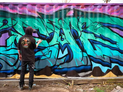 A street artist wearing a black hoodie and cap stands before an expansive graffiti mural, spray can in hand, adding details to the dynamic composition. The wall explodes with electric turquoise and deep blue wildstyle lettering, accented by magenta and purple geometric patterns that dance across the upper section. Set against the urban backdrop of Northeast Killingsworth Street in Portland's Alberta district, scattered debris and weathered concrete ground the scene in authentic street art culture.