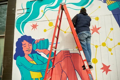 Artist Anke Gladnick climbs a red aluminum ladder to reach the upper portions of her vibrant mural "Let's Talk" on the exterior wall of Open Signal in Portland's Eliot neighborhood. The work-in-progress reveals her signature style of layered analog and digital elements, featuring flowing turquoise forms, geometric constellations of yellow dots, coral-pink star bursts, and a serene female figure with dark curls holding what appears to be a green water vessel. Gladnick, dressed in a black puffer jacket and jeans, stretches upward with concentrated focus, embodying the physical dedication required to transform architectural surfaces into dreamlike narratives.