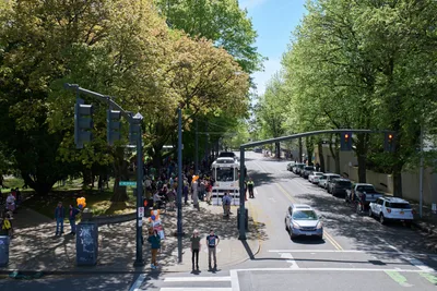 Crowds gather around a vintage white TriMet MAX Type 1 light rail car during its farewell ceremony at Holladay Park in Portland's Lloyd District. Dappled sunlight filters through mature spring foliage, casting dancing shadows across the assembled transit enthusiasts and officials in safety vests. The iconic train sits beneath traffic signals on the tree-lined street, marking the end of an era for Portland's original light rail fleet. Orange balloons and ceremonial touches add warmth to this bittersweet urban transit milestone.