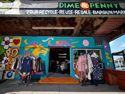 A vibrant thrift store facade on Northeast 15th Avenue in Portland's Alberta district showcases exuberant street art murals in turquoise, orange, and purple hues. Clothing racks spill onto the sidewalk beneath weathered wooden eaves, while colorful garments hang in the storefront windows. The hand-painted signage declares this a "recycle-reuse-resale-bargain mart," embodying the neighborhood's creative spirit and sustainable commerce ethos.