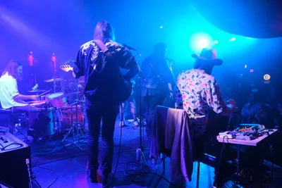 Roselit Bone commands the intimate stage at Dante's in Portland, bathed in electric blue and purple stage lighting that transforms the venue into a sonic cathedral. The lead performer, wearing a distinctive floral-patterned jacket and black hat, leans into their microphone while bandmates create a wall of sound behind them - a drummer in white and additional musicians shrouded in the moody atmospheric lighting. The cramped stage setup and tangled cables speak to the raw, underground energy of Portland's live music scene.