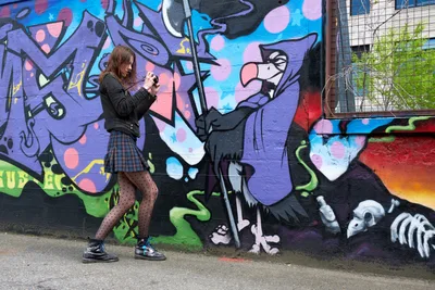 A young woman in plaid skirt and polka dot tights photographs a vibrant mural featuring cartoon-like characters in purple, pink, and blue hues along Southeast 3rd Avenue. The kaleidoscopic street art creates a dynamic backdrop as she crouches with her camera, capturing the creative energy of Portland's Hosford-Abernethy neighborhood. Spring light filters through the scene, illuminating both the detailed spray-painted imagery and the photographer's focused documentation of this urban canvas.
