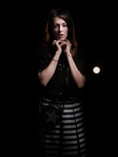 Rhianna Atwood emerges from deep shadows in this dramatic portrait captured in Portland's historic Kerns neighborhood. The chiaroscuro lighting sculpts her contemplative expression as she poses with hands clasped near her chin, wearing a black leather corset paired with a geometric striped skirt. A single warm light source pierces the darkness behind her, creating an intimate atmosphere of urban sophistication against the inky void.