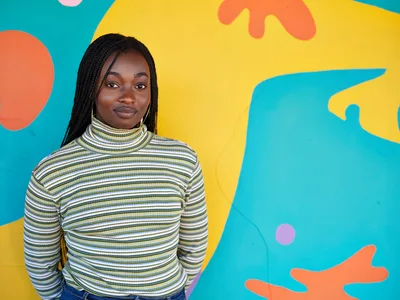 Naomi Likayi stands confidently before her completed mural in Portland's Alberta Arts District, wearing a striped turtleneck that echoes the rhythmic patterns of her artistic work. The explosive backdrop of turquoise, golden yellow, coral, and lavender organic shapes creates a dynamic canvas that transforms the Northeast Portland streetscape. Her serene expression and poised stance reflect the satisfaction of artistic completion, while the bold, flowing forms behind her pulse with contemporary energy against the urban setting.
