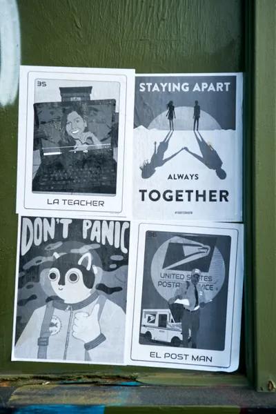Four photocopied flyers create a makeshift gallery on weathered olive-green wood in Portland's Concordia neighborhood. The black and white prints capture pandemic zeitgeist through humor and hope: a smiling teacher from a Polaroid labeled "LA TEACHER," silhouettes maintaining distance under "STAYING APART ALWAYS TOGETHER," a cartoon astronaut proclaiming "DON'T PANIC," and "EL POST MAN" featuring a postal worker and truck. The grassroots display transforms ordinary street surface into community commentary during COVID-19 lockdown.