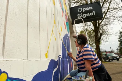 Maria Rodriguez, known as Sparkykneecap, methodically applies blue paint to her vibrant mural "Let's Talk" on the white exterior wall of Open Signal in Portland's Eliot neighborhood. Wearing wireless headphones and a striped shirt, the Mexican-American artist works with focused concentration as afternoon light illuminates her colorful work-in-progress. The scene captures the intimate process of public art creation, with the artist's paint bucket and brushes arranged nearby as she brings themes of identity and culture to life on Northeast Martin Luther King Jr. Boulevard.
