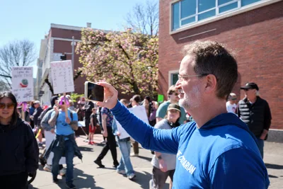A middle-aged man in a blue hoodie raises his smartphone to document the scene at a protest march in Portland's historic Old Town district. Behind him, demonstrators carry signs and gather on Southwest Ankeny Street, while spring cherry blossoms frame the brick buildings. The bright afternoon light illuminates the crowd as the man, identified as Tim Dickinson, captures the moment of civic engagement unfolding around him.