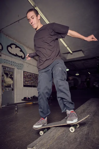 A focused skateboarder in muted earth tones navigates a weathered skateboard through the industrial interior of Commonwealth Skateboarding in Portland's Buckman neighborhood. Harsh fluorescent lighting cuts through the gritty atmosphere, casting stark shadows across concrete floors while vintage skate posters and graffiti-marked walls frame the scene. The rider's concentrated expression and fluid stance capture the meditative precision of street skating culture within this authentic underground retail space.